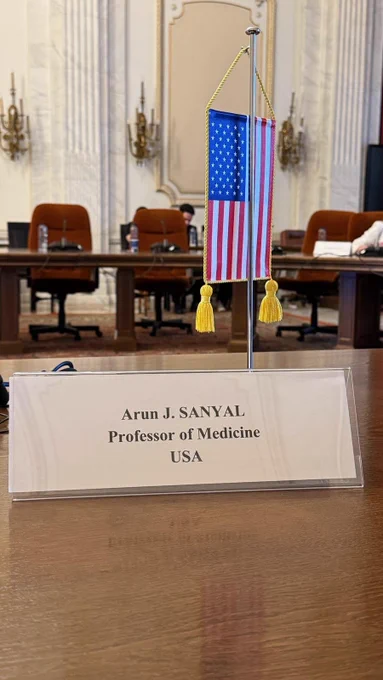 A conference room with a nameplate reading "Arun J. Sanyal, Professor of Medicine, USA" on a table. An American flag is placed next to the nameplate. Several chairs and water bottles are visible in the background.