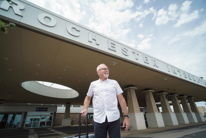 A smiling, elderly man stands outside Rochester International Airport with a rolling suitcase. He wears a white button-up shirt, dark gray slacks, black glasses, and a black watch.