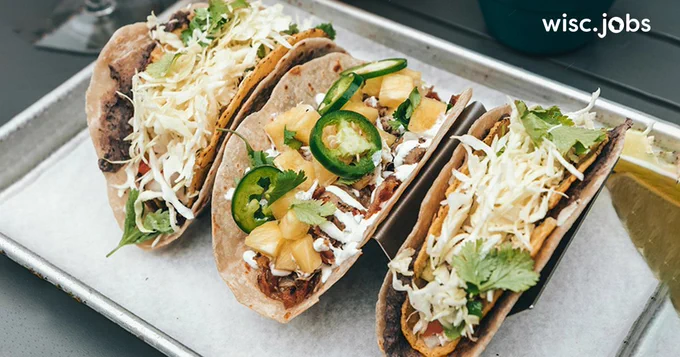Image: three soft tacos filled with shredded cabbage, pineapple chunks, jalapeño slices, cilantro, and drizzled with sauce on a metal tray; Wisc.Jobs