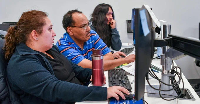 Three people seated side by side working on desktop computers in a modern office setting.
