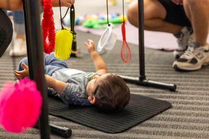 A child is lying on a black mat reaching up towards colorful hanging sensory toys, including a yellow brush and a red whisk. An adult kneels nearby, wearing athletic shoes.