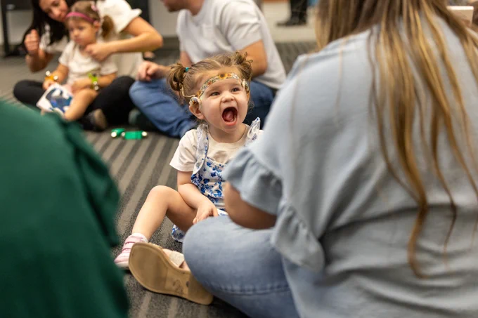 A child sitting on the floor with a joyful expression, surrounded by adults. The child's clothing is colorful, and the atmosphere seems lively and engaging.