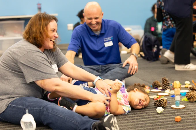 Two adults and a child are playing on a carpeted floor. The child, lying on their back, is laughing while the adults, also smiling, engage with them. The floor is scattered with pinecones and colorful play items. 