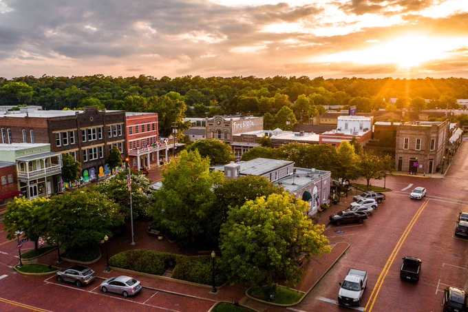 Aerial view of a charming town square at sunset, featuring historic brick buildings and neatly parked cars. The streets are paved with red bricks, and trees add greenery to the scene. The sun sets in the background, casting a warm glow over the area.
