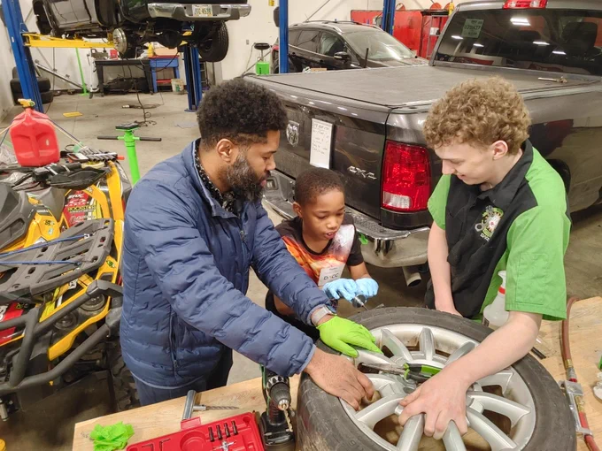 A man and two children work on a car's wheel in a garage, surrounded by tools and equipment.