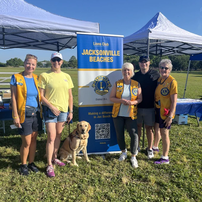Members of the Jacksonville Beaches Lions Club stand smiling in front of their blue and yellow club banner under event tents, with a yellow guide dog sitting at their feet on the grass at Jacksonville VisionWalk.