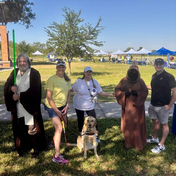 Five people pose on the grass at Jacksonville VisionWalk, including two in Star Wars costumes, two casually dressed participants, and a yellow guide dog sitting calmly at the front with tents and trees in the background.