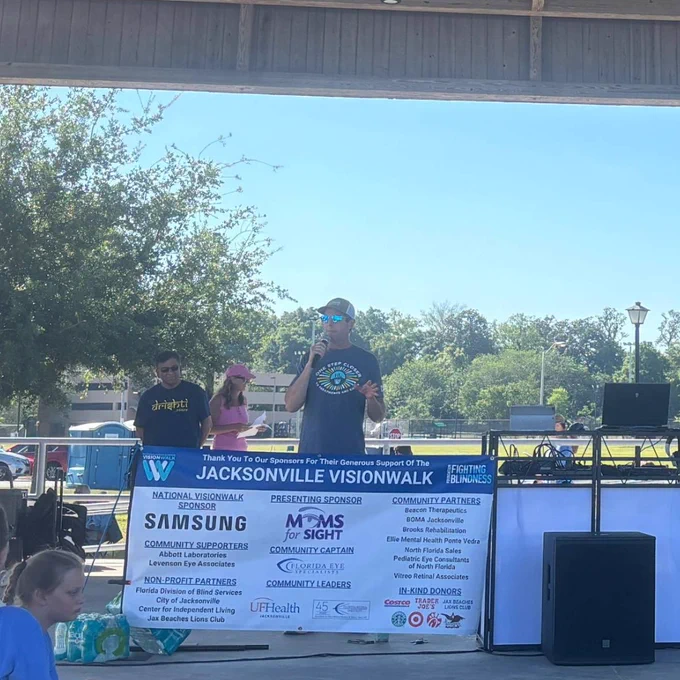 A speaker in a Jacksonville VisionWalk T‑shirt holds a microphone on an outdoor stage in front of a sponsor banner and sound equipment, with trees and a clear blue sky behind him.