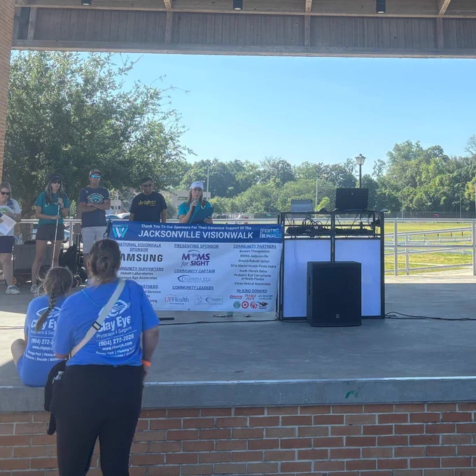 Speakers stand on an outdoor stage at Jacksonville VisionWalk behind a large sponsor banner, while two participants in blue event shirts watch from the front of the crowd on a sunny morning.