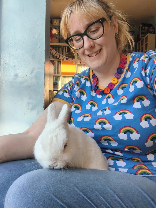 White Netherlands dwarf rabbit sat on a (his owner) lady's knee, he's looking down towards them. The lady is wearing blue jeans, a blue t shirt with rainbows on it, a button necklace, green glasses and has blonde hair. The lady is looking at him and smiling.