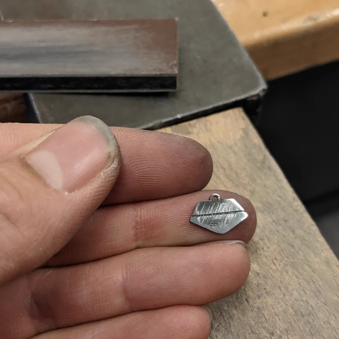 Fingertips holding a tiny meteorite capsule charm with the Widmanstätten pattern etched across its flat surface.