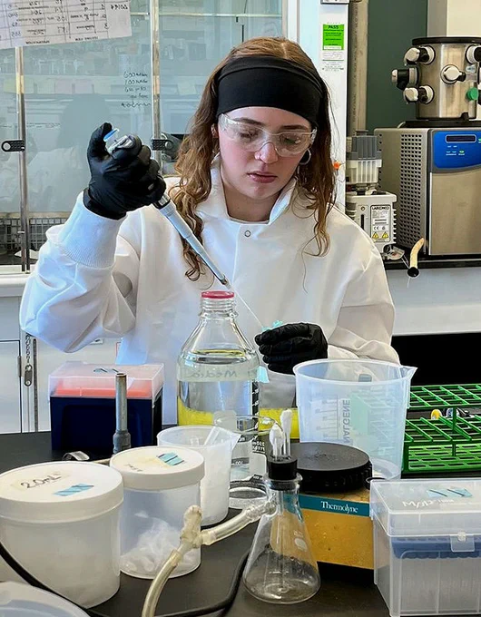 Summer internship student working in a biotech lab. She's wearing a lab coat and protective goggles.