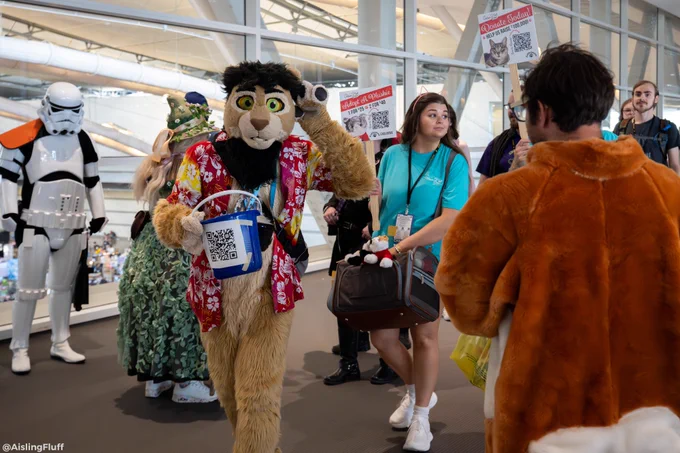 A lion fursuit wearing a red hawaiian shirt with a blue bucket for the Anthrocon Charity Bucket Brigade as various attendees walk around him