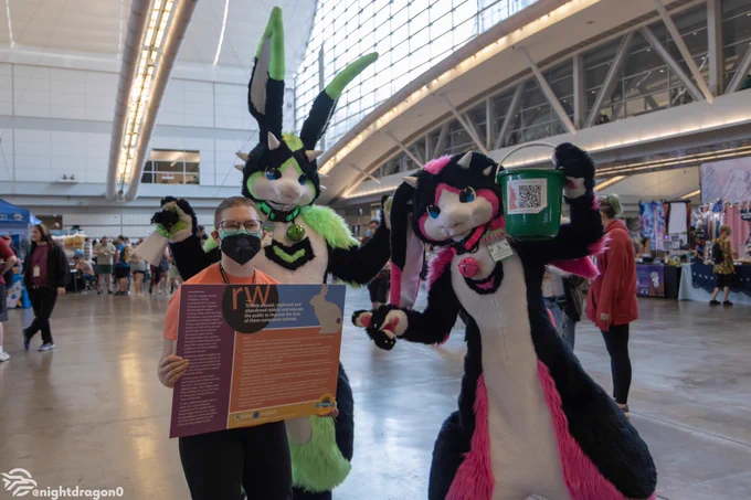 A pair of bunnies, green/black and pink/black respectively, with a green charity bucket helping to raise money for the Anthrocon 2023 charity.