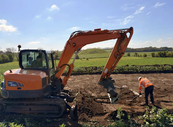 A large orange mechanical digger digs soil@in the foreground, while an archaeologist in orange hi-vis shovels nearby. Skipsea castle mound in the background, visible through the arch of the diggger arm.