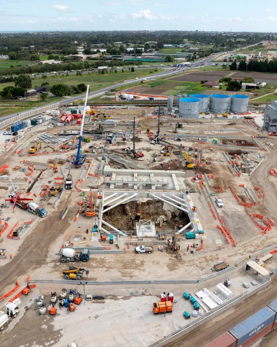 Aerial view of a large construction site with cranes and various construction vehicles. There are multiple structures in progress, surrounded by orange barriers. Four large blue cylindrical tanks are positioned towards the top right. A highway and some greenery are visible in the background.