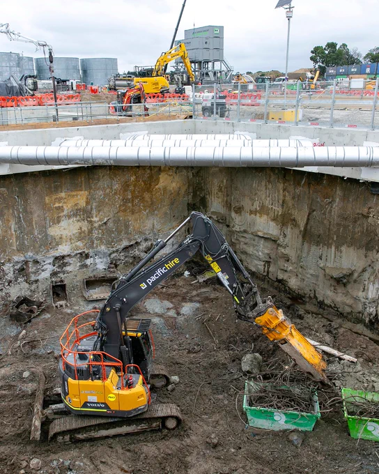 Construction site featuring a large pit with an excavator labeled "PacificHire" working inside. In the background, additional machinery and equipment are visible, surrounded by safety barriers and industrial structures.