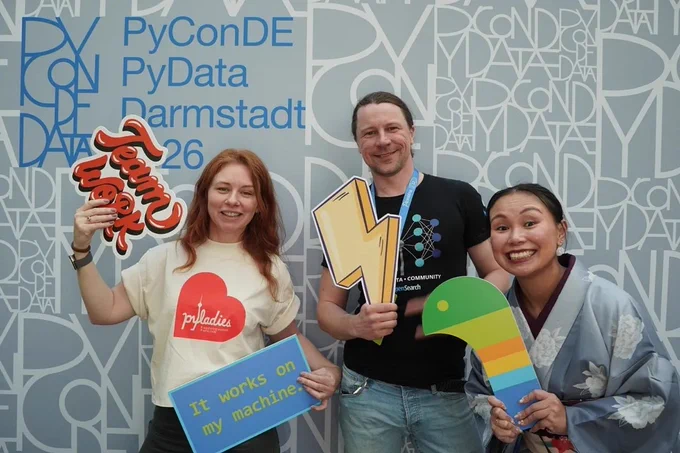 Three individuals pose with colorful signs at PyConDE 2026 in Darmstadt, showcasing team spirit and tech enthusiasm.