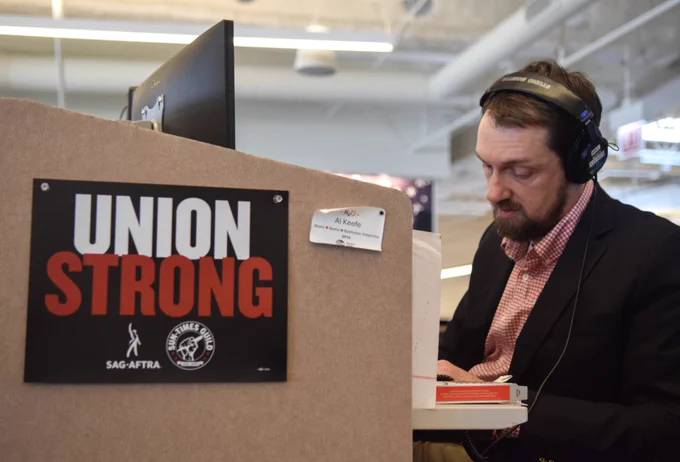 SAG-AFTRA member and WBEZ community engagement editor Al Keefe works at his desk with a sign that says “UNION STRONG” with both SAG-AFTRA and the Sun-Times Guild logo on it pinned to the side.