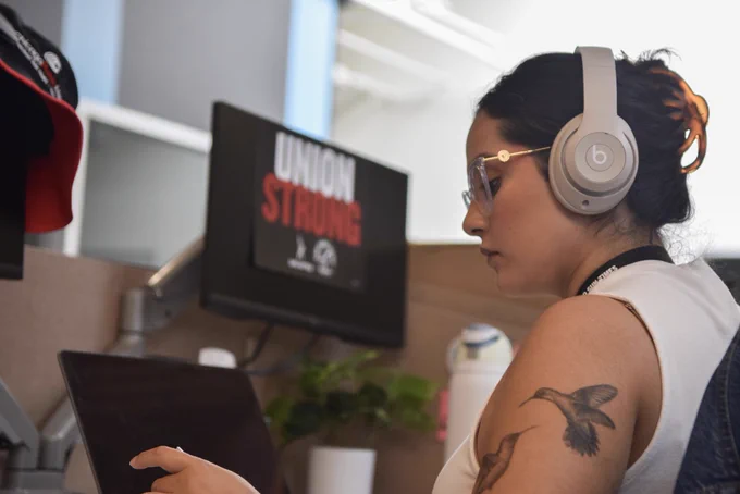 Sun-Times Guild member and reporter Cindy Hernandez works at her desk with a sign over a second computer monitor that says “UNION STRONG” with both the SAG-AFTRA and the Sun-Times Guild logos on it.