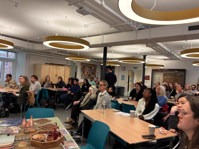 Group of people sitting and listening at the session that Turquoise Mountain hosted at the Museum of Oxford for the Marmalade Festival (UK) in partnership with the Skoll World Forum. They are sitting on blue chairs and by large, wooden tables. [Photo by Turquoise Mountain]