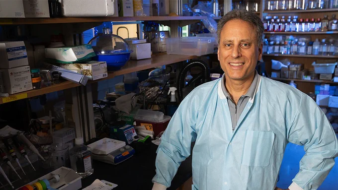 A man with gray hair stands in a lab while wearing a light blue protective lab coat over a gray button-up shirt. Behind him are a lab bench and shelves filled with scientific equipment.