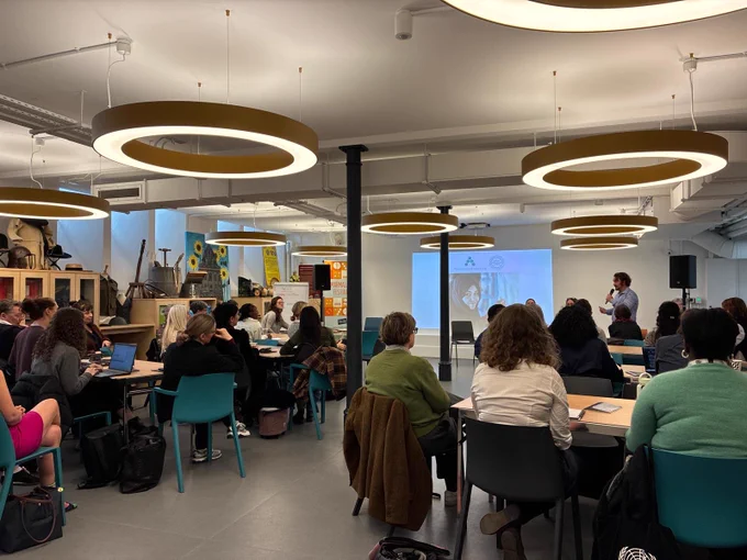 A group of people sitting on blue chairs and listening to a speaker, who is standing in front of the room. The speaker is speaking into a microphone with two people sitting on chairs next to him. Behind them is a projection of a photo of a smiling woman. [Photo by Turquoise Mountain]