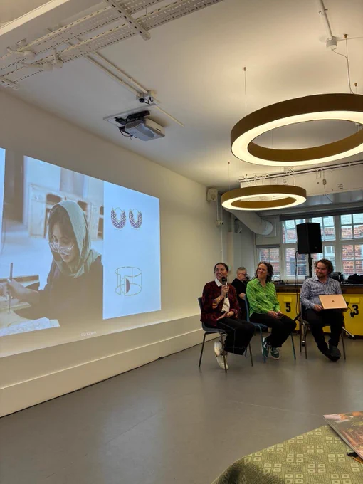 Three people sitting on chairs and looking at a projection of a woman, who is doing calligraphy or drawing. One of the people is speaking into a microphone. [Photo by Turquoise Mountain]