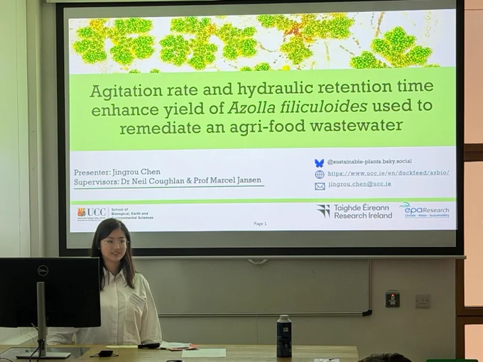 Jingrou Chen is standing beside a large presentation screen in front of the hall at ENVIRON 2026. The title of the presentation is on the screen: "Agitation rate and hydraulic retention time enhance yield of Azolla Filiculoides used to remediate and agri-food wastewater.