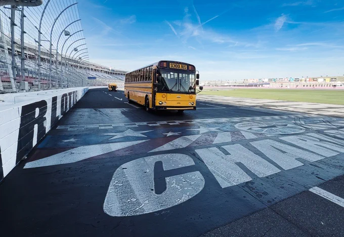 A Thomas school bus on the track at Charlotte Motor Speedway, with its characteristic yellow color standing out against the asphalt. Another bus follows in the distance, and the speedway's grandstands are visible under a clear blue sky.