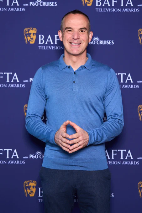 Martin Lewis CBE is stood smilng towards the camera with a light blue long-sleeved top on. He is holding his hands towards each other in front of him. There is a dark blue step and repeat board behind him with the BAFTA Television Awards with P&O Cruises logo on it. 