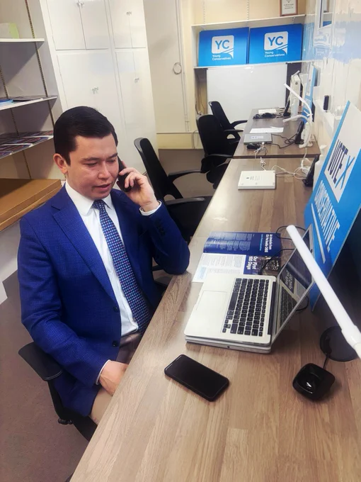 A man in a blue suit sitting at a desk, speaking on a phone. 
