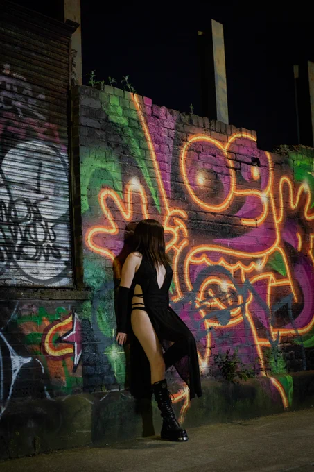 Trans Princess Jezebel stands in the street at night in a flowing black cloak and knee high leather New Rock boots leaning against a wall with a large colourful neon graffiti mural of Bart Simpson with his tongue out.