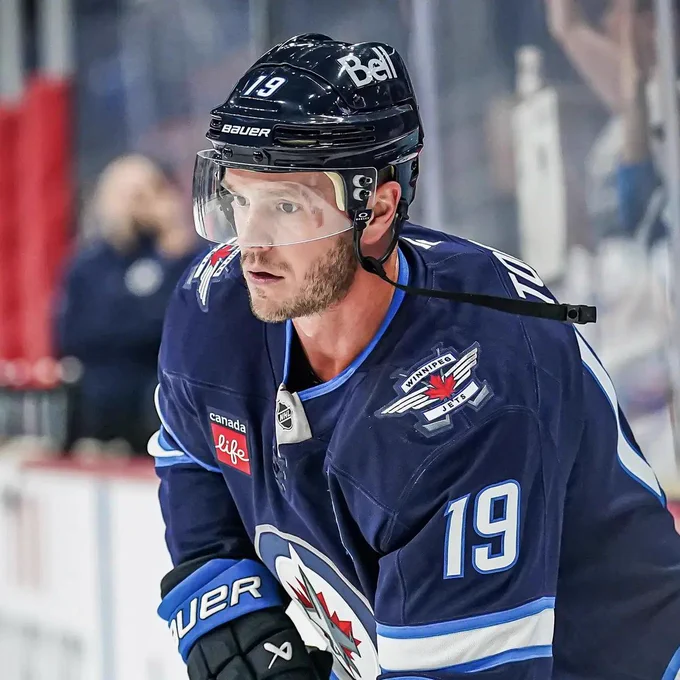 Jonathan Toews wearing a dark blue Winnipeg Jets jersey with number 19 and a captain’s patch, along with a black helmet with a clear visor and Bauer branding. A Bell logo is visible on the helmet and a Canada Life patch on the jersey. He is leaning forward on the ice, looking ahead with a focused expression, with rink boards and blurred spectators in the background.
