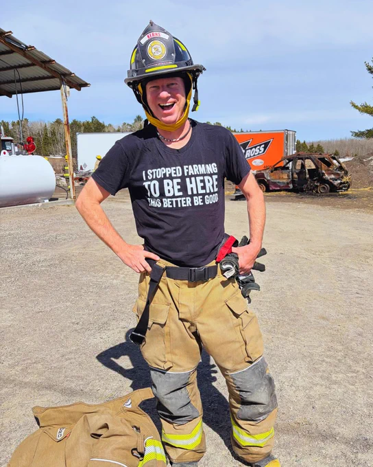 A person wearing firefighter gear and a helmet stands outdoors, smiling. Their shirt reads, "I stopped farming to be here. This better be good." In the background, there is a building, a large storage tank, and a red trailer labeled "Ross." The setting appears to be a rural or industrial area under a clear sky.