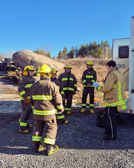 A group of firefighters in yellow helmets and reflective suits stands together outdoors near a large rock. One firefighter, dressed similarly with additional Canadian flag patches, appears to be speaking to the group. The scene includes a fire truck and a rocky, forested background under a clear blue sky.