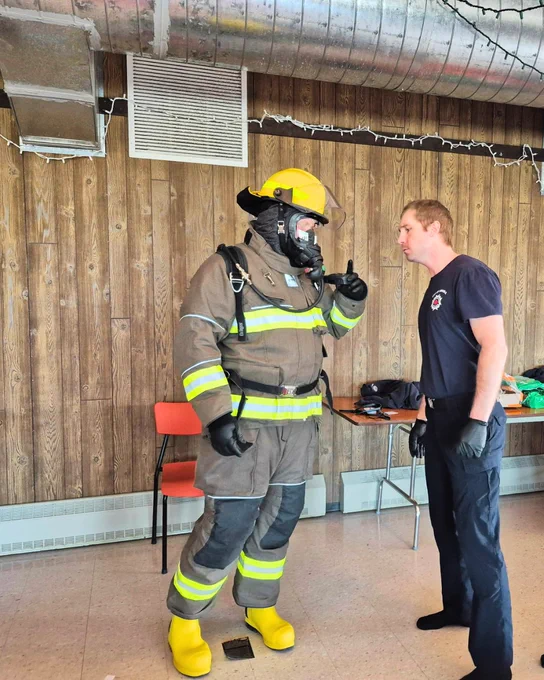 A person in full firefighter gear, including a helmet and mask, stands indoors next to a person in a dark uniform. The room has wood-paneled walls and a chair with equipment on a table nearby.