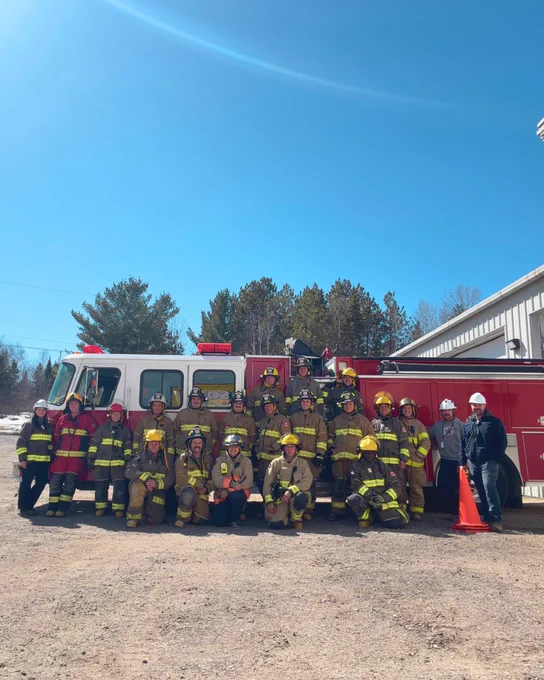 A group of firefighters in uniform and trainers poses in front of a red fire truck on a sunny day, with trees in the background. A few people are wearing white helmets on the right side of the group.