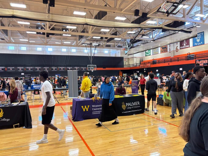 Students and representatives interact at various college booths inside a gymnasium during a college fair event.