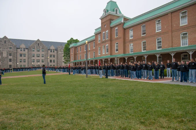 Cadets wearing black shirts and blue jeans stand in formation on a grassy quad in front of a long brick building with a covered walkway.