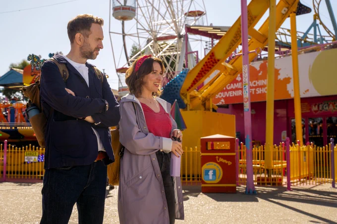 From Trying. Rafe Spall as Jason and Esther Smith as Nikki walk through an amusement park together. Jason gives Nikki a skeptical look as she chatters happily.