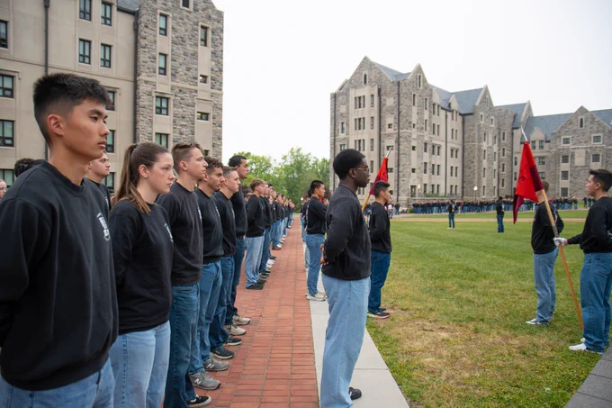 Cadets in black shirts and blue jeans stand in multiple rows along a brick path on a grassy quad, with campus residence halls visible in the background.