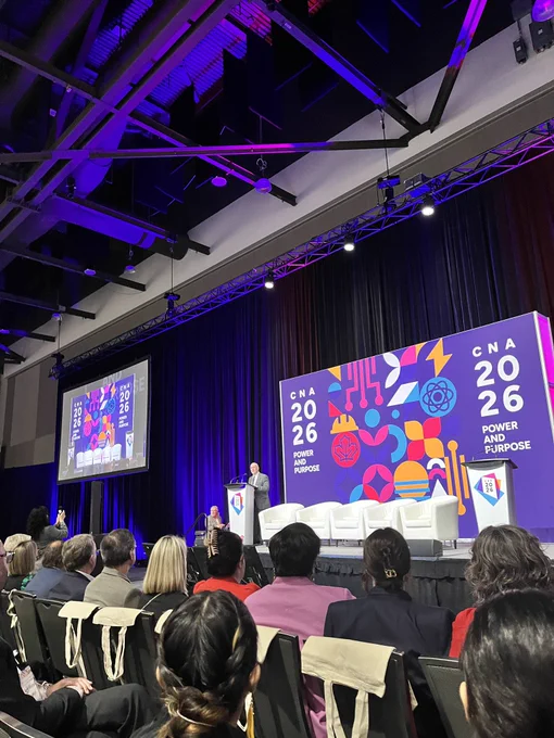 A man stands on a conference stage with 4 white arm chairs next to him, behind is a screen with a colourful graphic. On the graphic says CNA 2026