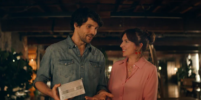 From Trying. Colin Morgan as Kerry wearing a chambray shirt and Esther Smith as Nikki wearing a pink shirt smile while engaged in a conversation with one another.