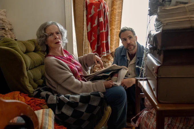 From Trying. Rafe Spall as Jason and Paula Wilcox as Sandra Ross are seated next to each other surrounded by papers and suitcases. Paula is holding a newspaper in her hands.