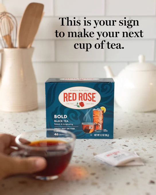 Box of Red Rose Bold Black Tea on a kitchen counter with a hand holding a glass cup of tea in the foreground with a saying "this is your sign to make your next cup of tea."