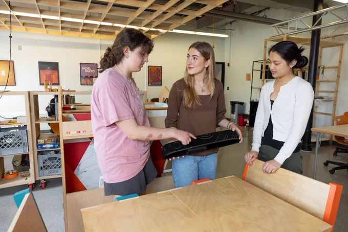 Three people are in a workshop setting, discussing a rectangular object. They appear engaged and are surrounded by wooden furniture and shelving. The background features posters. The environment suggests a creative or collaborative workspace.
