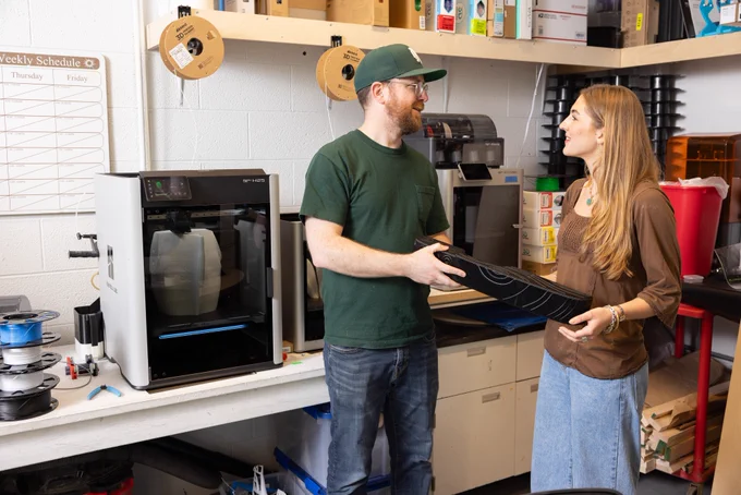 Two people stand in a workshop with 3D printers and spools of filament. One person holds a black object resembling a large gear. Shelves and a weekly schedule are visible in the background.