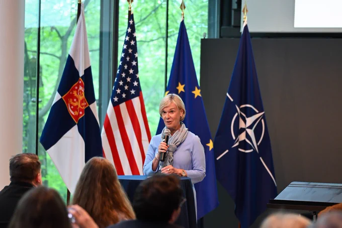 Ambassador in light blue speaking to an audience in front of flags.