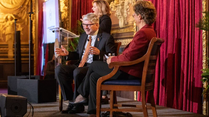 A man in a suit gestures as he sits in a chair on a stage with a woman in a red suit